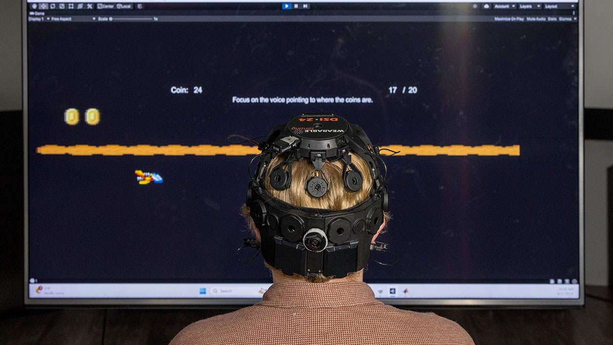 person wearing a cap with electrodes on it looks at a monitor displaying an orange line.