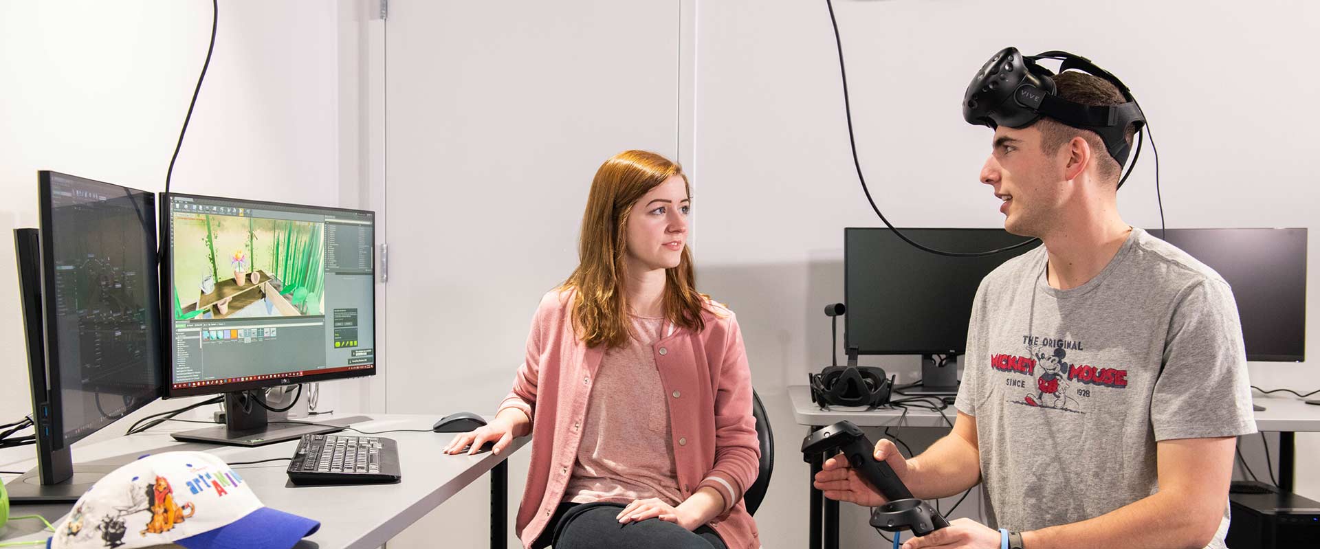 two students talking, with one wearing a virtual reality headset and holding handhelds.