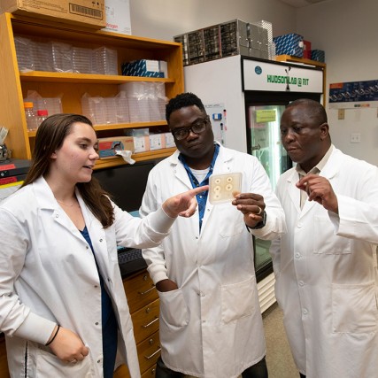 two professors and student wearing lab coats looking at bacteria sample.