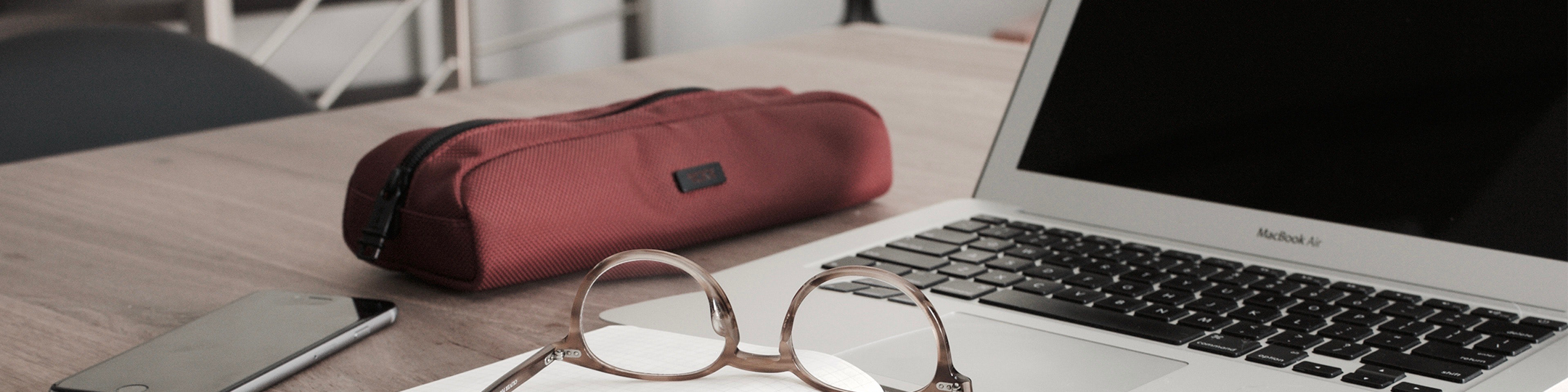 angled photo of a laptop, glasses, phone, and a red pencil holder on a desk