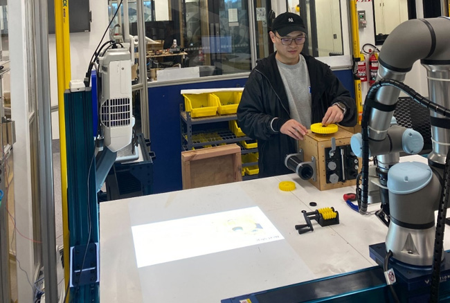 Person standing at an assembly bench with cameras and projector.