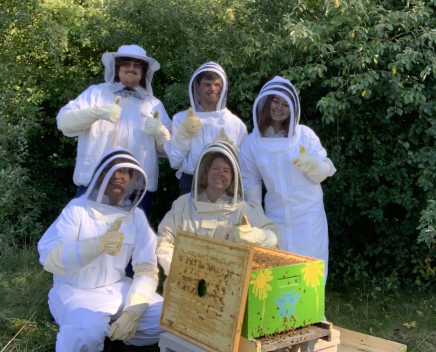 A photo of five members of the RIT Beekeeping Club Bee Board. They are standing aroudn an open yellow and green bee hive and giving a thumbs up.