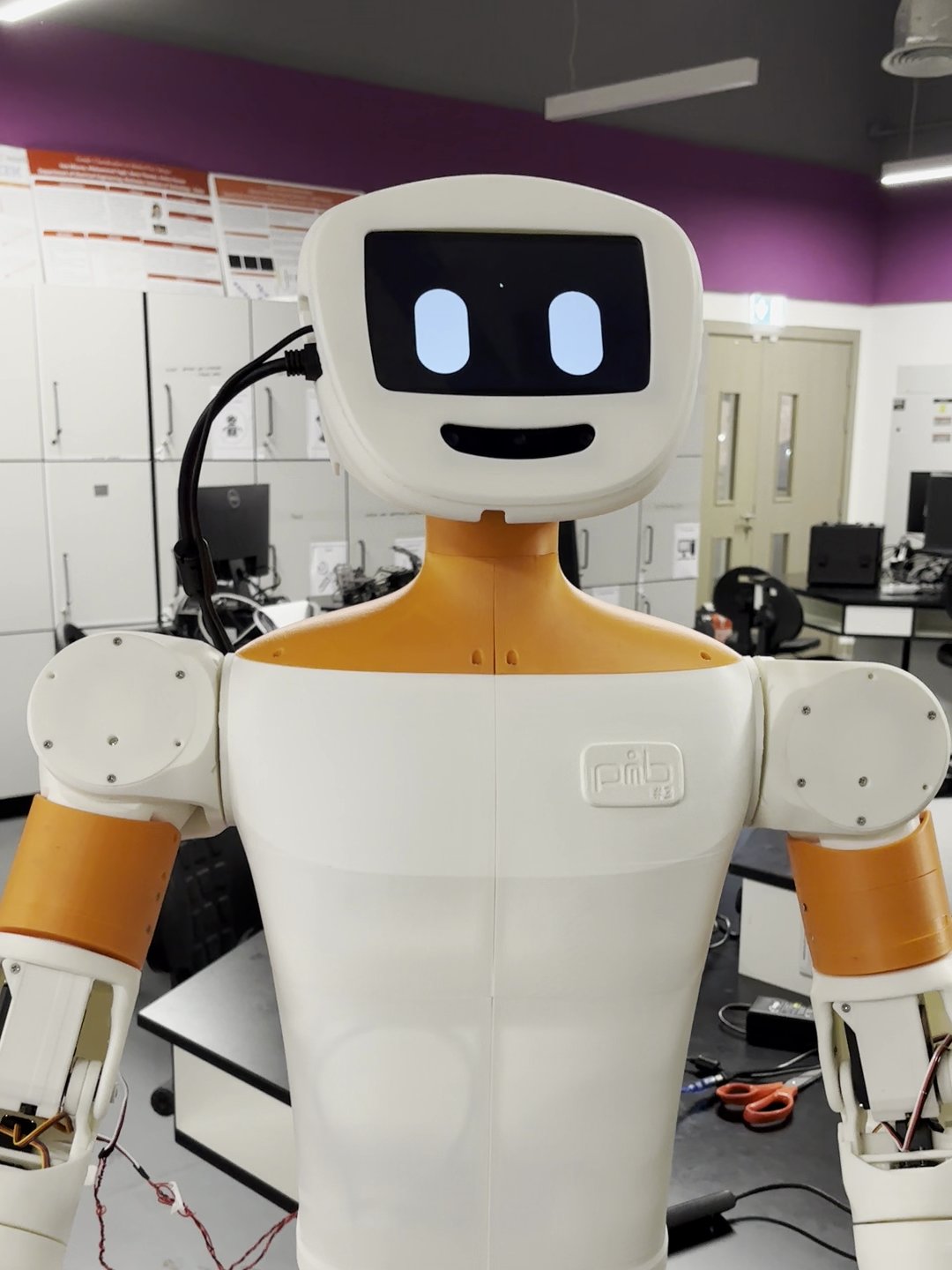 A white and orange humanoid social robot stands in a laboratory setting, featuring a digital display screen, articulated arms, and visible technical equipment in the background.