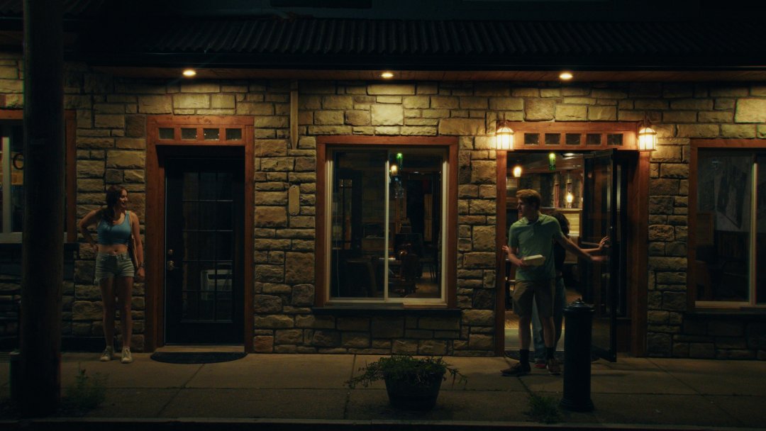 A woman stands outside a stone building. A man and another woman exit the door of the building.