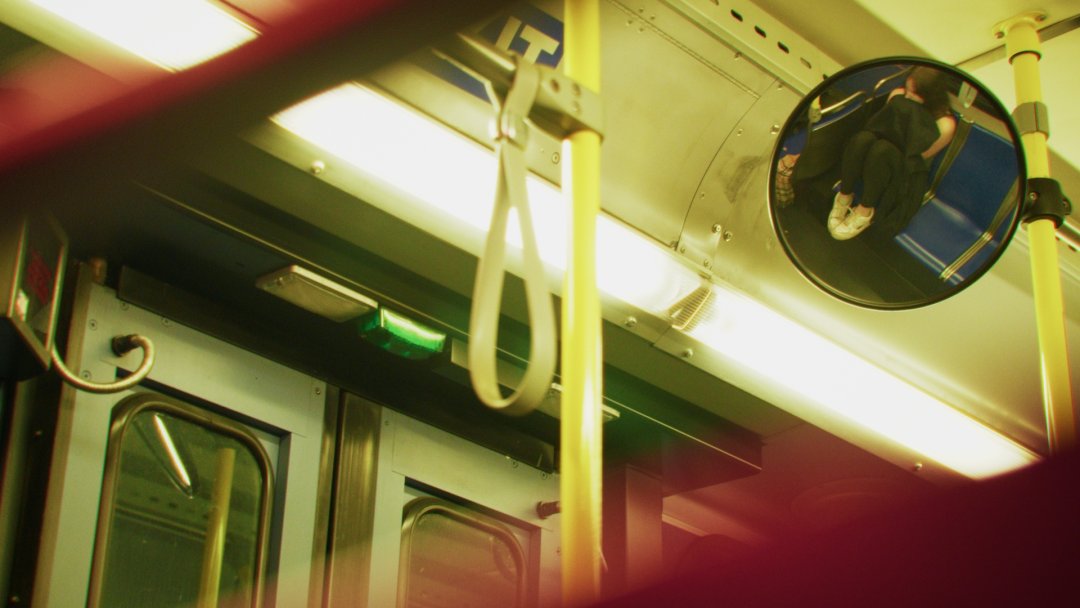 A woman is framed in a circular bus mirror. She is curled up on the seat of the bus.