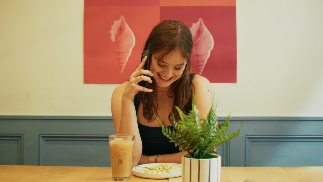 A woman smiles, holding a phone to her ear, in a bright cafe. A red poster with pictures of conch shells hangs behind her.
