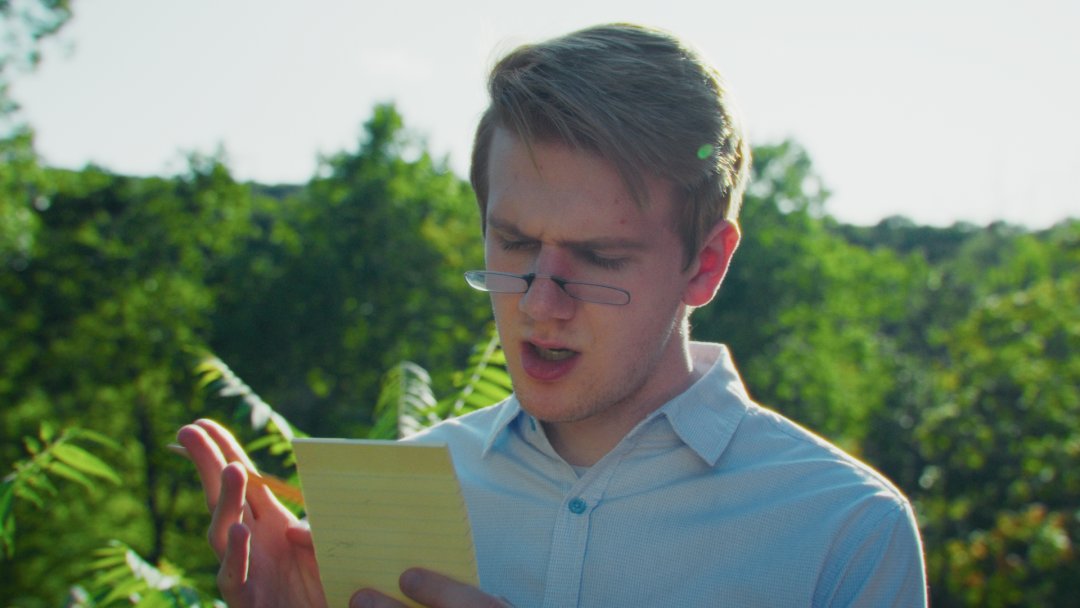 A man with small reading glasses stands in front of foliage, holding a notepad