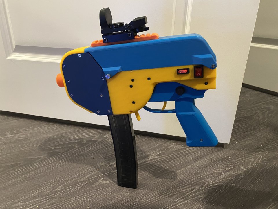 A medium sized blaster constructed from yellow, blue and navy filament. It is balanced on the ground using its mag. A small sight is attached to the top.
