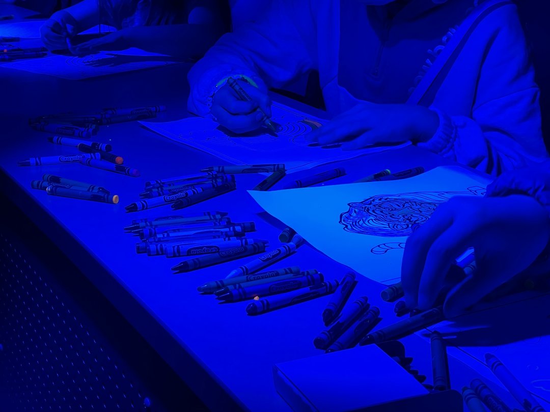 Several people are doing a coloring activity at a table. Their hands, some coloring pages, and many crayons are visible, all lit with a very intense blue light that makes it difficult to discern the crayon colors.