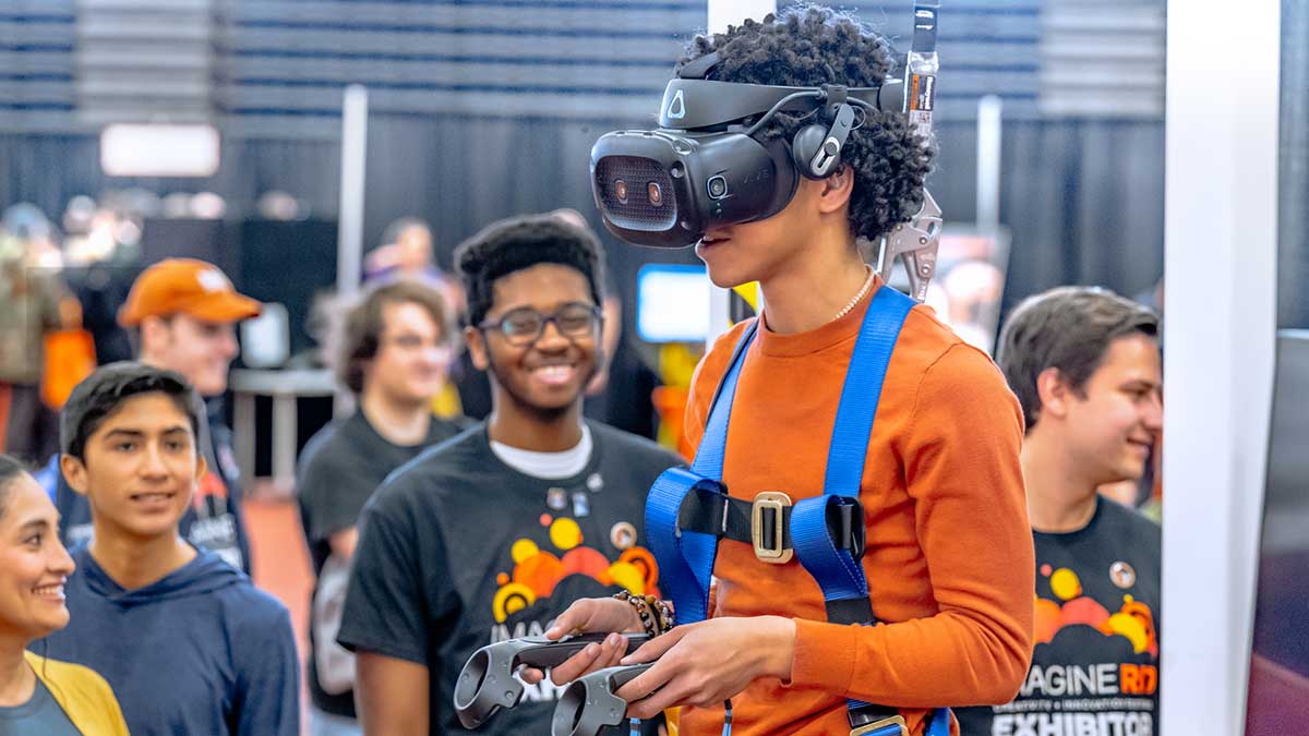 A young man wearing a VR headset and a motion-tracking harness interacts with a virtual reality exhibit at Imagine RIT.
