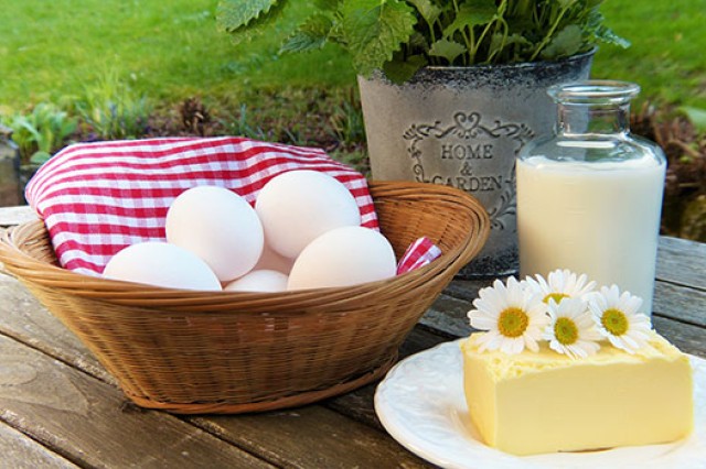 An artistic photo of eggs in a basket, milk in a glass bottle, and a large block of cheese with daisies on top of it.