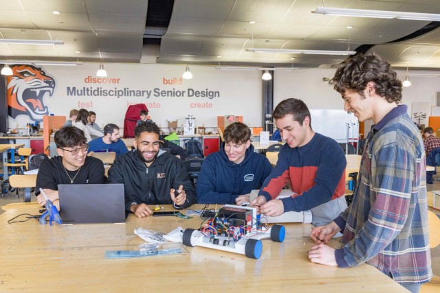 a group of students sit at a wooden table in a workshop, one works on a laptop while the others work on a robot car