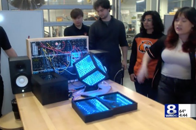 students stand at a table looking at a lit up cube connected to a computer panel.