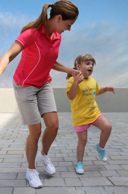 Adult helping a young girl skip, both lifting one leg playfully on a paved outdoor area.