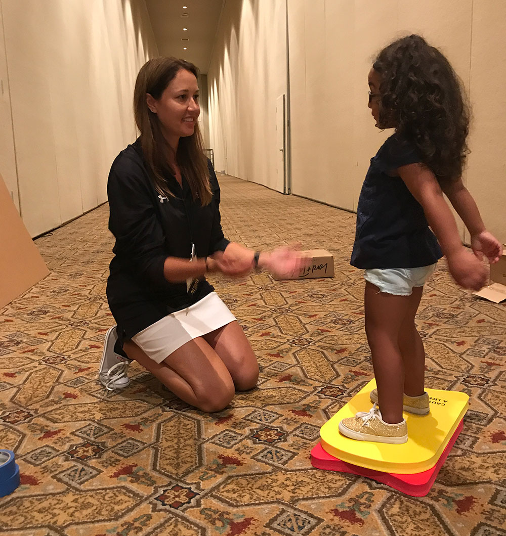 Adult kneels to engage with a young girl balancing on colorful foam pads in a hallway.