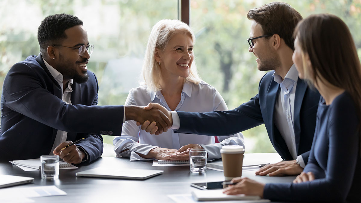 A group of business professionals sit at a table while two people shake hands and smile during a meeting.