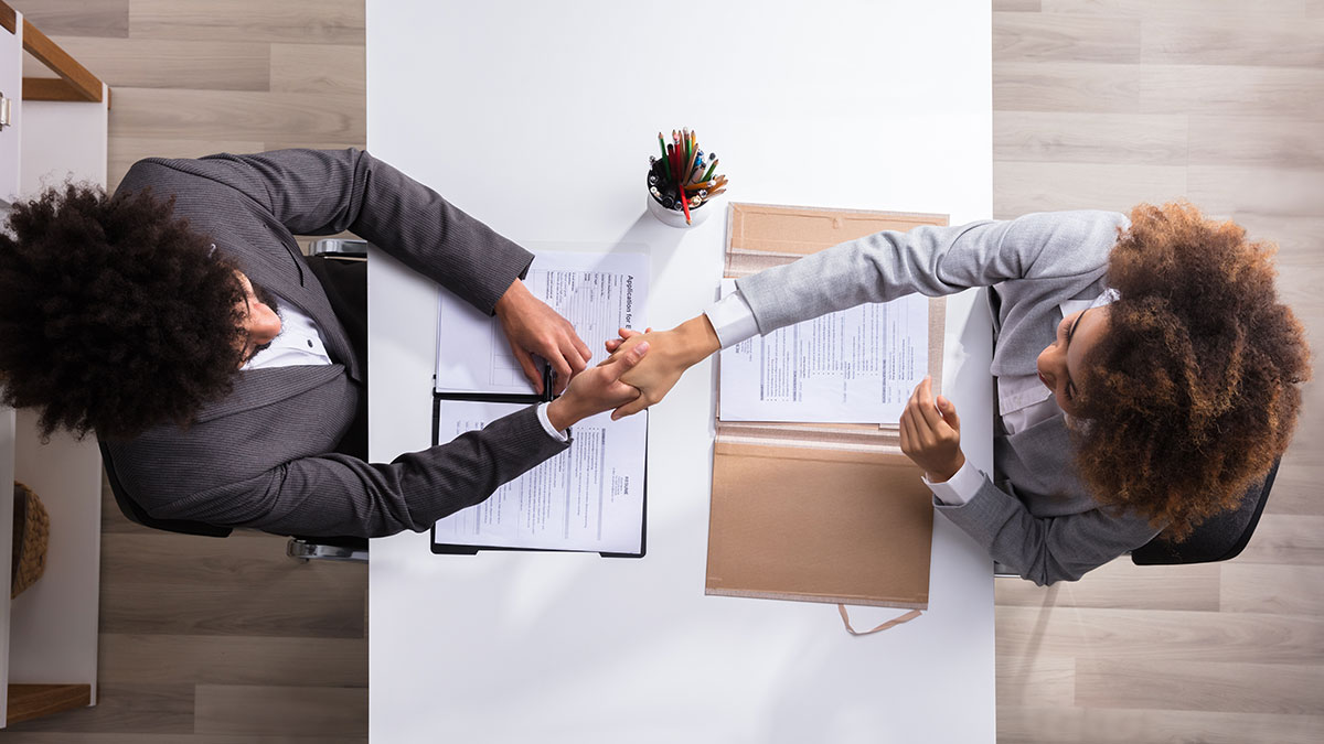 Two professionals seated across a desk shake hands over documents during a meeting.