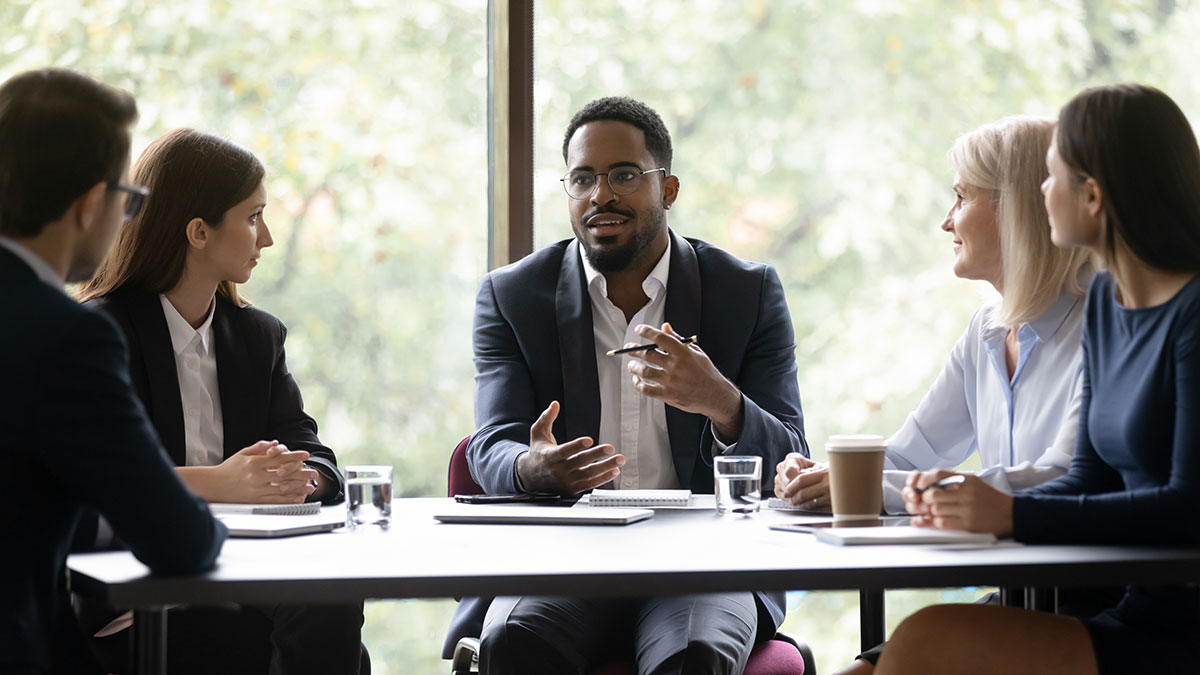 A business professional speaks to colleagues during a meeting at a conference table.