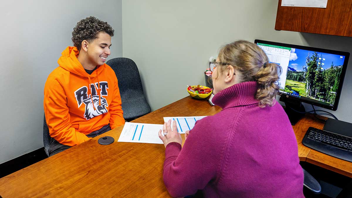 A student wearing an R I T hoodie discusses course options with an advisor in a small office setting.