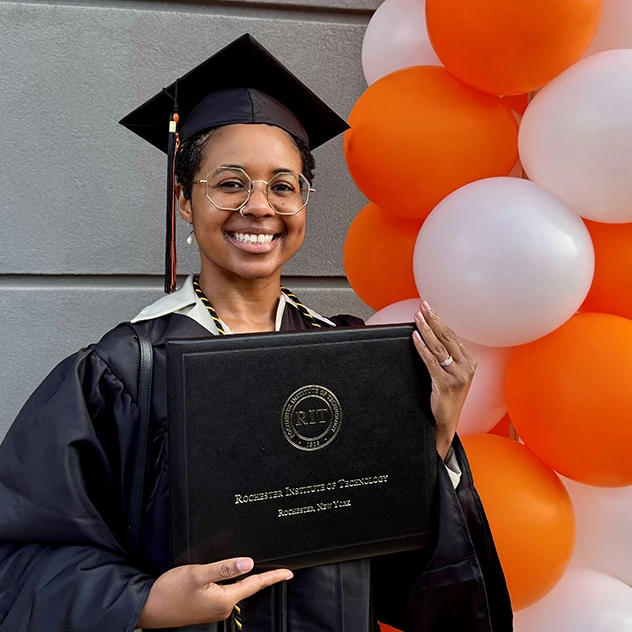 Person in cap and gown holding an RIT diploma cover, smiling beside orange and white balloons.