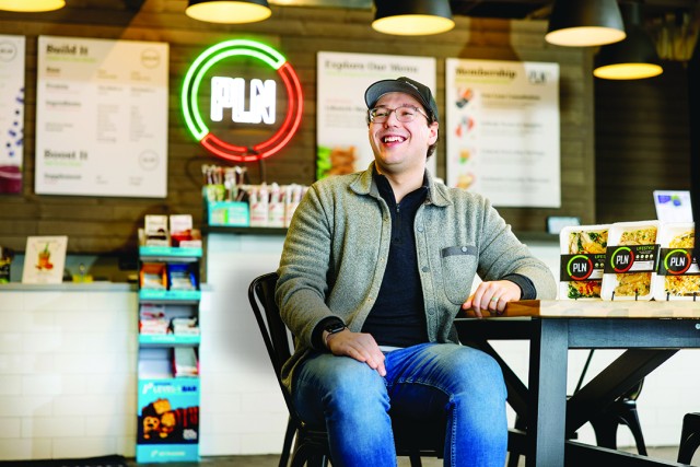 a man in a gray zip up shirt and baseball cap sits at a table in a casual industrial restaurant space.