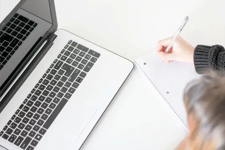 Top view of a person writing something in a notebook with a laptop in front of them.