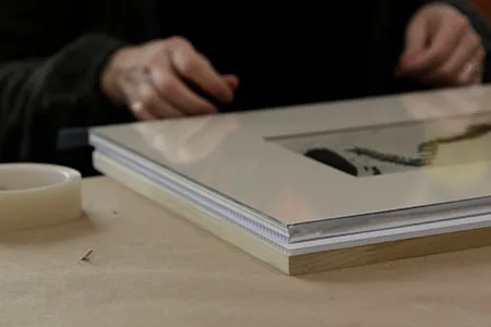A photo in the process of being framed on a table. It has a piece of glass on the front and a backing behind it. There is a person sitting at the table working on framing the photo.