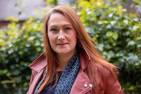 Photo of Dr. Emma Richardson standing in front of a bush outside. She has red hair and is wearing a red jacket.