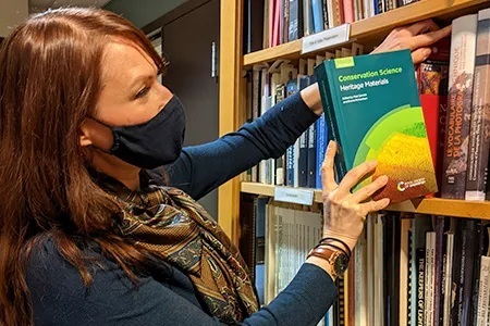 Photo of a woman wearing a mask grabbing a book titled "Conservation Science Heritage Materials" from a bookshelf.