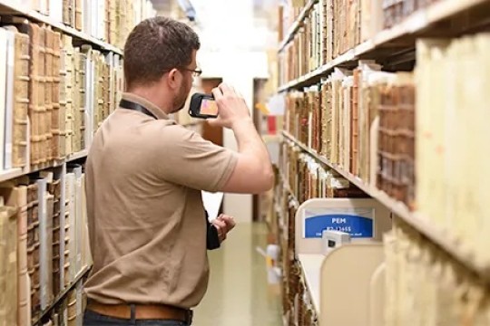 Man standing between two rows of books with his back to us. He is looking through a camera.