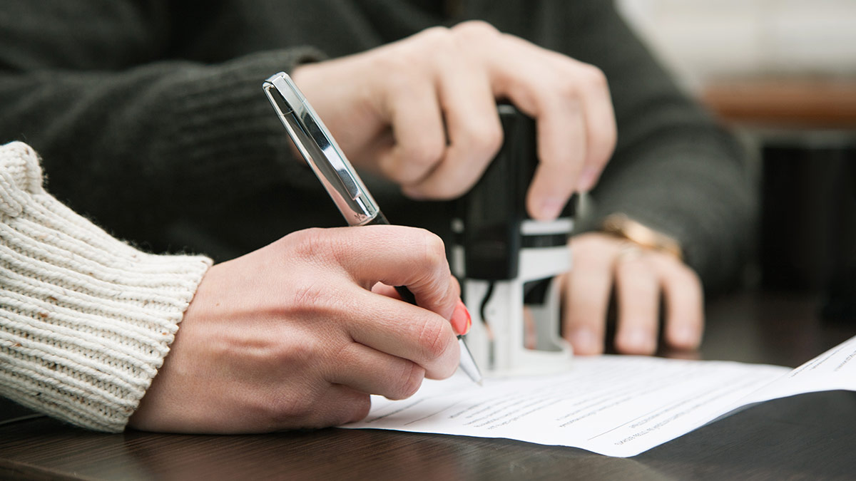 A close up of a person signing a document while another person stamps it.
