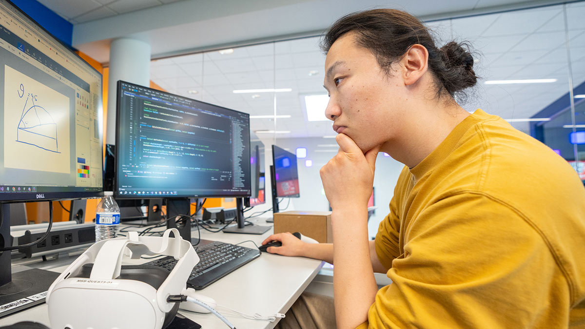 a student looks at two computer monitors, with a V R headset on the deskin front of him.