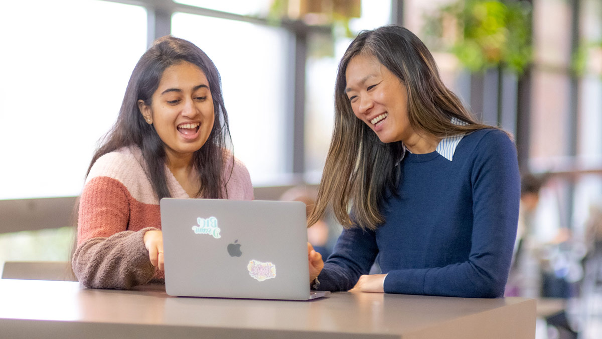 two college students smiling while looking at a laptop.