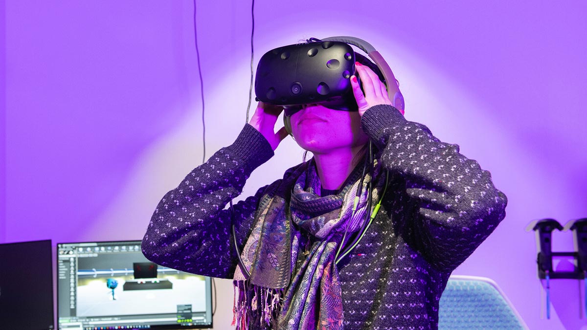 college student holds V R goggles to her head as she looks around, with a purple hue in the background.