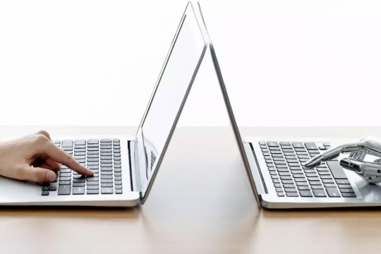 two laptops back to back on a table with a human hand typing on one and a robot hand typing on the other