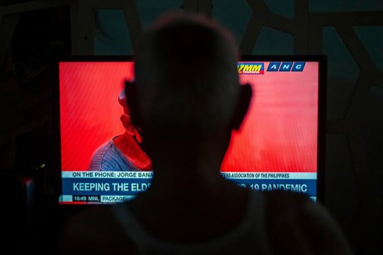 An elderly person watches a television news broadcast discussing the impact of the COVID-19 pandemic on older adults, with a red-toned image of a masked individual on the screen.