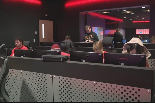 Students in a dimly lit esports or cybersecurity lab, working at computer stations with red LED lighting. A man wearing an 'RIT' hoodie stands behind them.