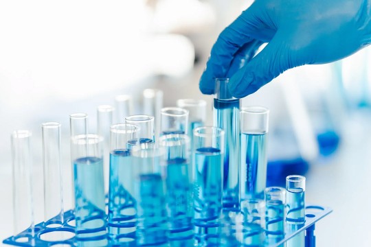 Gloved hand holding a test tube filled with blue liquid among multiple test tubes in a laboratory setting.
