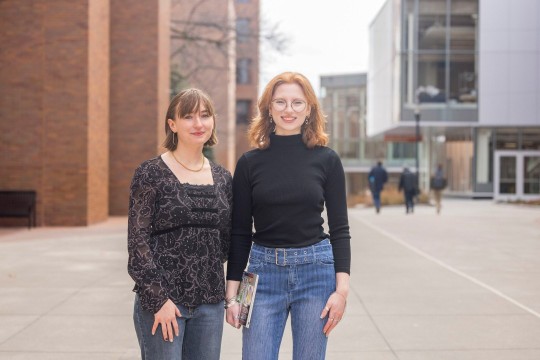Two young women stand side by side on a college campus, smiling at the camera. One wears a black lace top, while the other wears a black turtleneck and jeans, holding a book in her hand.