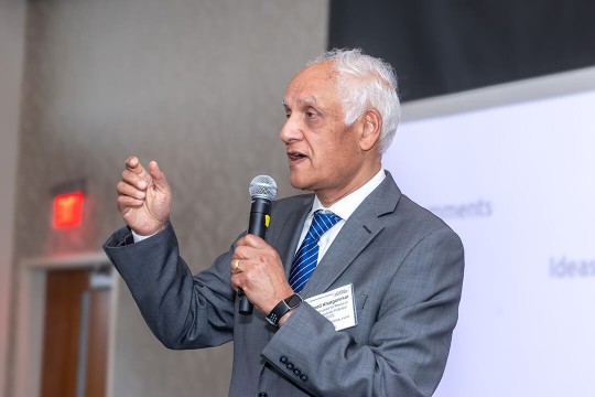 Pramod Khargonekar stands in grey suit and blue tie with microphone addressing audience