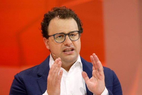 Man with glasses and small microphone speaks in front of a red background.