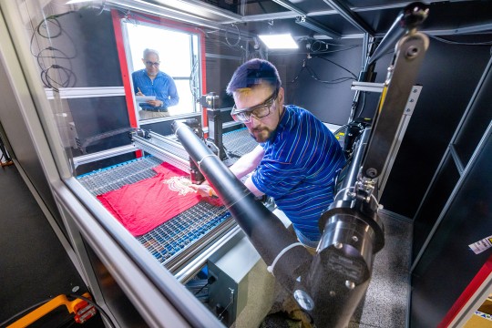 A man wearing safety goggles works with a t-shirt on a large machine while another man looks on