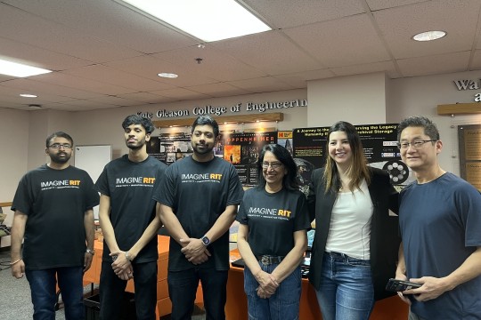 Six people pose for a photo in front of an academic poster. People are wearing shirts that read Imagine RIT.
