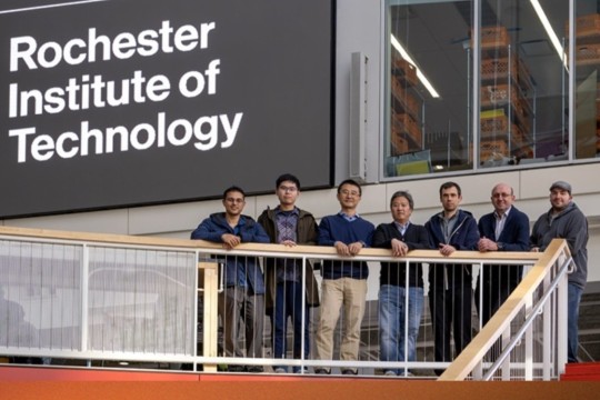 Seven people stand by the railing in the RIT SHED with a Rochester Institute of Technology sign behind them.