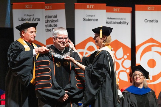 A man and woman put a silver commemorative adornment on William H. Sanders on a stage.