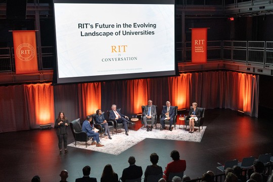 six people sit in chairs on a stage for a panel. a screen behind them reads R I T in conversation.