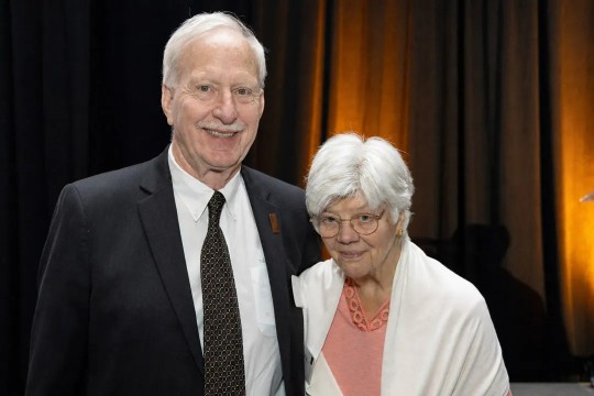 Older couple in suit and dress clothes stand in front of an orange backdrop. 