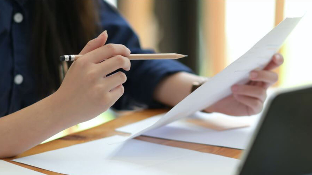 Person holding a pencil while reviewing a sheet of paper at a desk, with additional papers laid out nearby in a well-lit indoor setting.