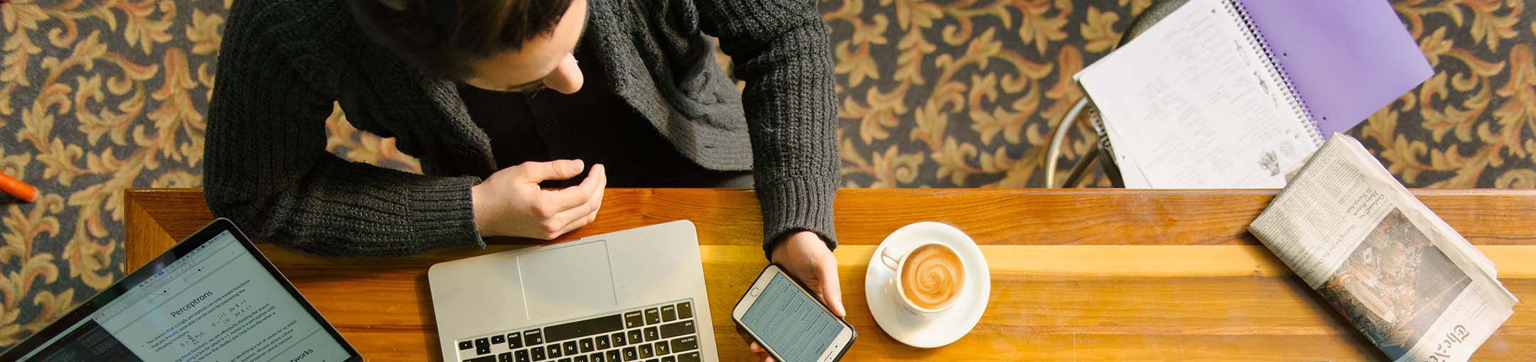 Overhead view of a person sitting at a table with a laptop, cup of coffee, cell phone and tablet.