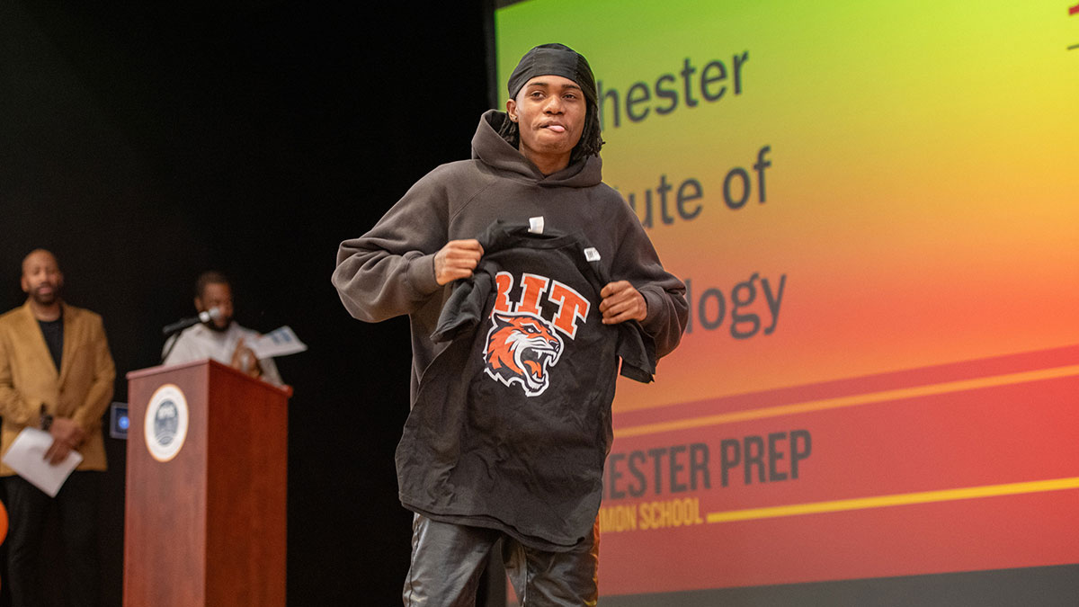 a high school student holds an R I T T-shirt while walking across a stage.